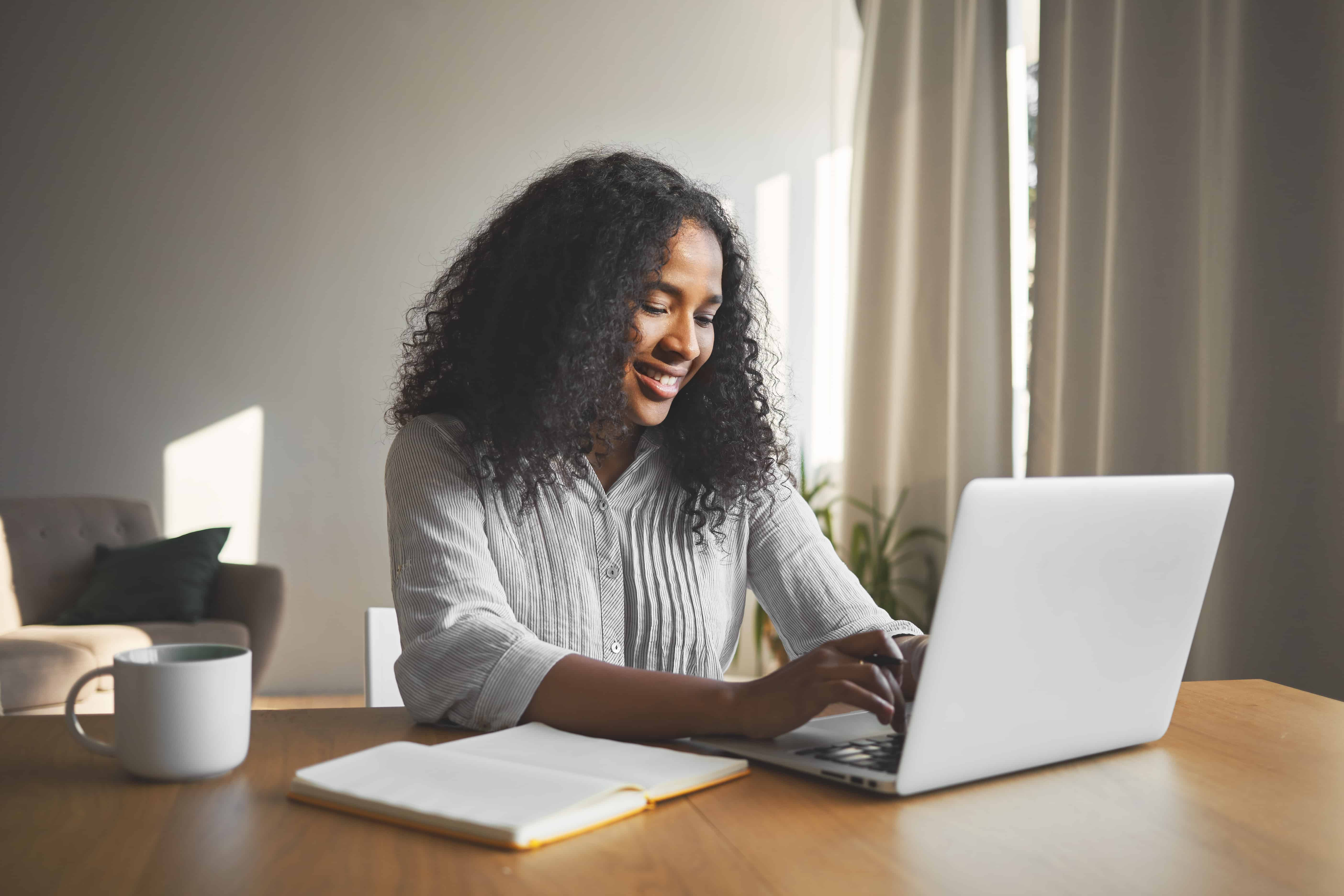 gorgeous positive young dark skinned female blogger keyboarding generic laptop smiling being inspired while creating new content her travel blog sitting desk with diary mug 1